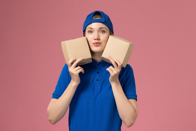 Corriere femminile di vista frontale in capo uniforme blu che tiene i piccoli pacchetti dell'alimento di consegna sulla parete rosa, lavoro dipendente di servizio di consegna