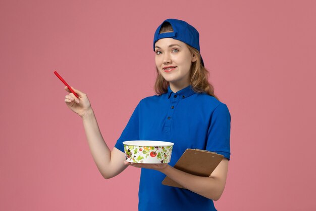 Corriere femminile di vista frontale in capo uniforme blu che tiene la ciotola di consegna e la penna del blocco note sulla parete rosa-chiaro, impiegato di consegna di servizio di lavoro