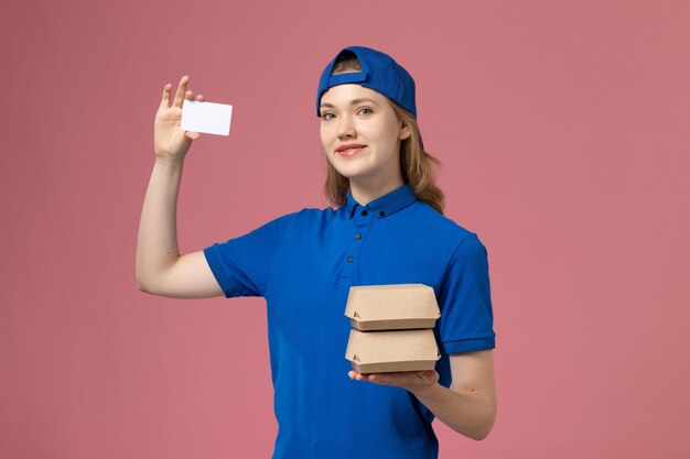 Corriere femminile di vista frontale in capo uniforme blu che tiene piccoli pacchetti di cibo di consegna e carta sull'impiegato di consegna di servizio del fondo rosa