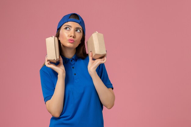 Corriere femminile di vista frontale in capo uniforme blu che tiene piccoli pacchi di consegna pensando sulla parete rosa, consegna dell'azienda di servizio dei dipendenti