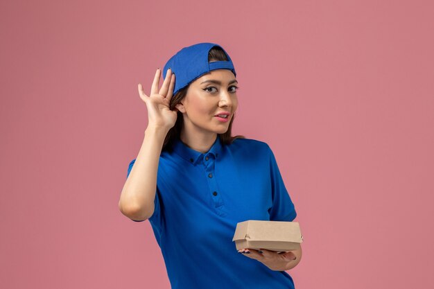 Corriere femminile di vista frontale in capo uniforme blu che tiene piccolo pacchetto di consegna che prova a sentire sulla parete rosa, lavoratore di lavoro di consegna di servizio del dipendente