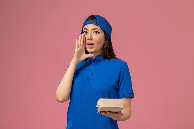 Corriere femminile di vista frontale in capo uniforme blu che tiene piccolo pacchetto di consegna e che chiama sul muro rosa, consegna del servizio dei dipendenti