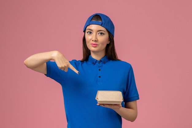 Corriere femminile di vista frontale in capo uniforme blu che tiene piccolo pacchetto di consegna sulla parete rosa, lavoratore di lavoro di lavoro di consegna di servizio di impiegato