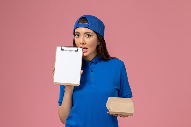 Corriere femminile di vista frontale in capo uniforme blu che tiene piccolo pacchetto di consegna vuoto con il blocco note sulla parete rosa, consegna dell'azienda di servizio dei dipendenti di lavoro