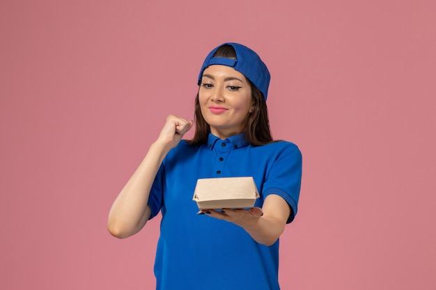 Corriere femminile di vista frontale in capo uniforme blu che tiene piccolo pacchetto di consegna vuoto e che si rallegra sulla parete rosa, consegna dell'azienda di servizio dei dipendenti