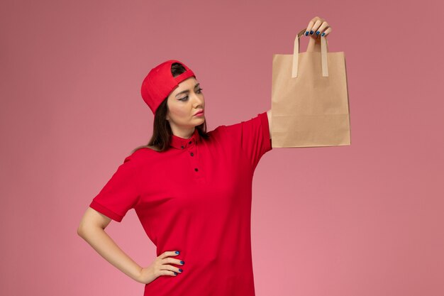 Corriere femminile di vista frontale in capo uniforme rosso con il pacchetto di carta di consegna sulle sue mani sulla parete rosa, impiegato di consegna del lavoratore uniforme