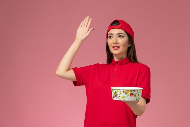 Corriere femminile di vista frontale in capo uniforme rosso con la ciotola di consegna sulle sue mani che fluttuano sulla parete rosa chiaro, impiegato di consegna di servizio