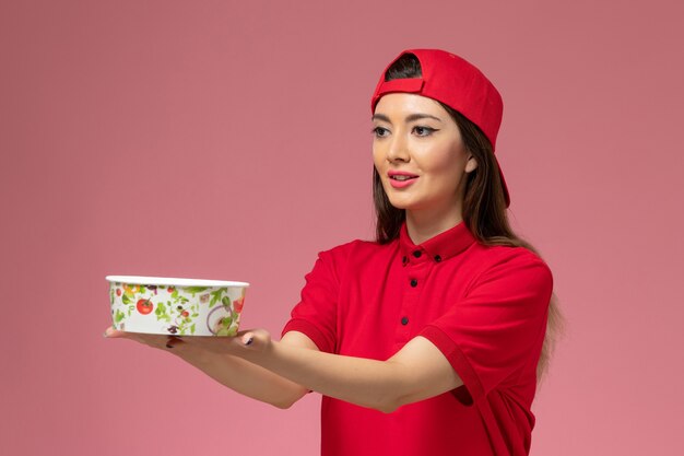 Corriere femminile di vista frontale in capo uniforme rosso con la ciotola di consegna sulle sue mani sulla parete rosa-chiaro, ragazza di lavoro degli impiegati di consegna di servizio