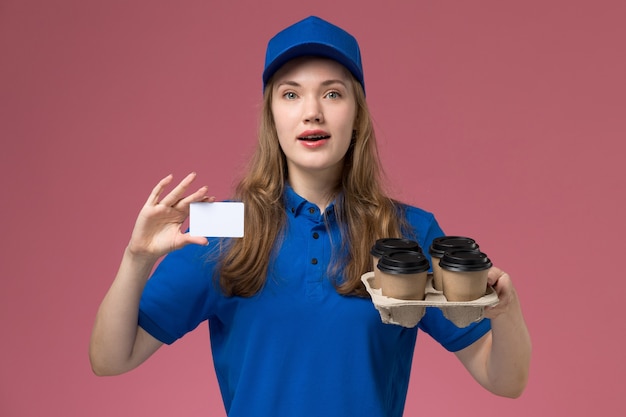 Corriere femminile di vista frontale in carta uniforme blu della tenuta e tazze di caffè marroni di consegna sul lavoratore dell'azienda uniforme di servizio scrivania rosa chiaro