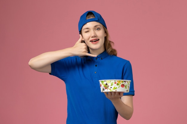 Corriere femminile di vista frontale in mantello blu uniforme che tiene la ciotola di consegna sulla parete rosa, ragazza dell'impiegato di consegna di servizio