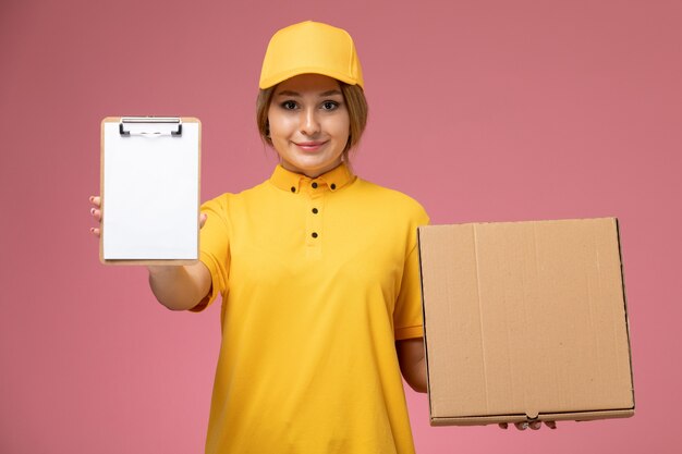 Corriere femminile di vista frontale in mantello giallo uniforme giallo che tiene la scatola di cibo del blocchetto per appunti sul colore femminile di consegna uniforme del pavimento rosa