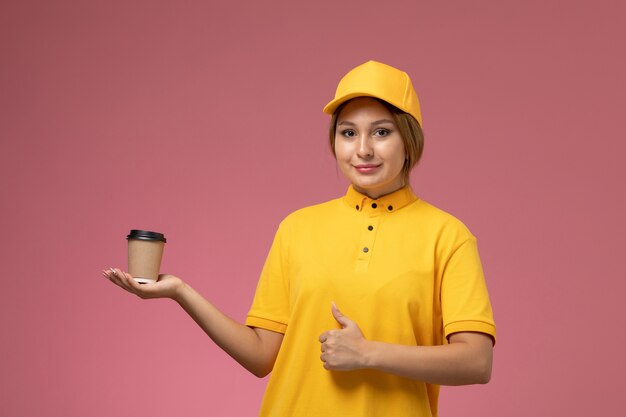 Corriere femminile di vista frontale in mantello giallo uniforme giallo che tiene la tazza di caffè marrone di plastica con il sorriso sul colore femminile di consegna dell'uniforme rosa della scrivania