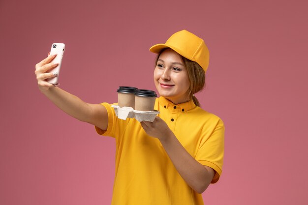 Corriere femminile di vista frontale in mantello giallo uniforme giallo che tiene le tazze di caffè che prendono un selfie sul colore di lavoro di lavoro di consegna uniforme di sfondo rosa