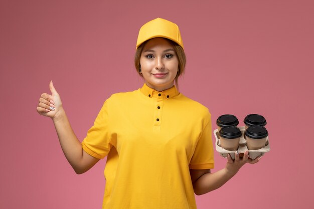 Corriere femminile di vista frontale in mantello giallo uniforme giallo che tiene le tazze di caffè di plastica con un leggero sorriso sul lavoro di colore del lavoro di consegna uniforme del fondo rosa