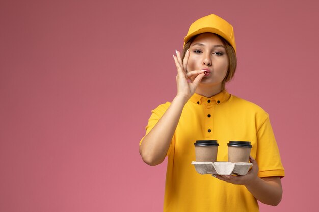 Corriere femminile di vista frontale in mantello giallo uniforme giallo che tiene le tazze di caffè marroni di plastica e che posa sul colore femminile di consegna dell'uniforme rosa della scrivania