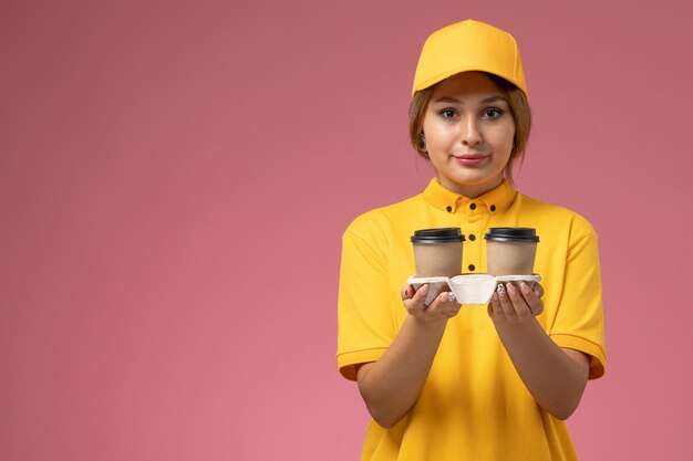 Corriere femminile di vista frontale in mantello giallo uniforme giallo che tiene le tazze di caffè marroni di plastica sul colore del lavoro di consegna uniforme della scrivania rosa