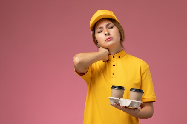 Corriere femminile di vista frontale in mantello giallo uniforme giallo che tiene tazze di caffè con mal di collo sul lavoro di lavoro di consegna uniforme sfondo rosa