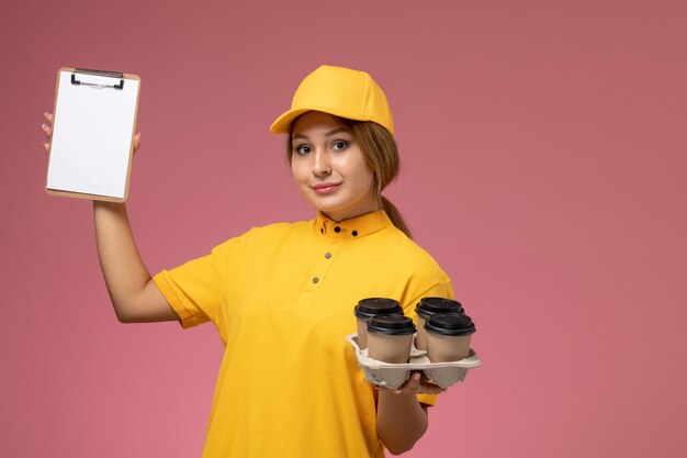 Corriere femminile di vista frontale in mantello giallo uniforme giallo che tiene tazze di caffè e blocco note sul colore di lavoro di lavoro di consegna uniforme di sfondo rosa