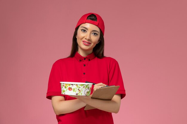 Corriere femminile di vista frontale in mantello rosso uniforme con blocco note rotondo ciotola di consegna sulle sue mani scrivere note sulla parete rosa chiaro, lavoro dipendente consegna uniforme