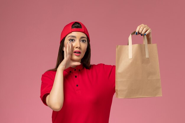 Corriere femminile di vista frontale in mantello rosso uniforme con pacco di carta di consegna sulle sue mani che bisbiglia sul muro rosa, impiegato di consegna di lavoro uniforme