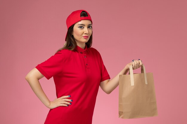 Corriere femminile di vista frontale in mantello rosso uniforme con pacco di carta di consegna sulle sue mani sul muro rosa chiaro, impiegato di consegna uniforme