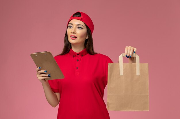 Corriere femminile di vista frontale in mantello rosso uniforme con pacco di consegna di carta e blocco note sulle sue mani sul muro rosa, impiegato di consegna uniforme