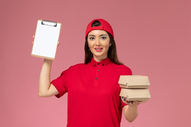 Corriere femminile di vista frontale in mantello rosso uniforme con piccolo pacchetto di cibo di consegna e blocco note sulle mani sul muro rosa chiaro, impiegato di consegna del servizio di lavoro