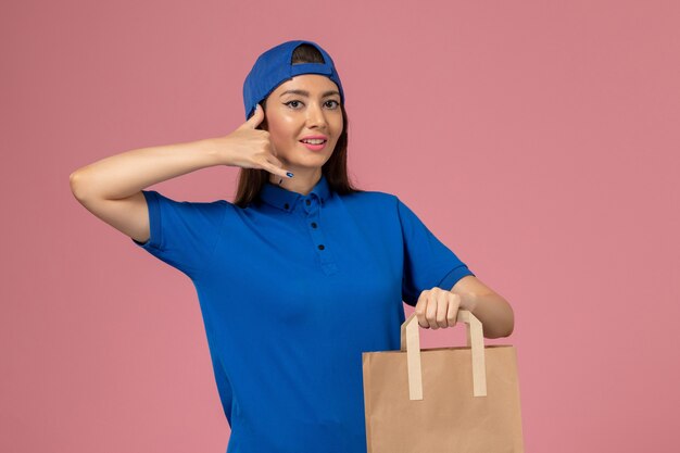 Corriere femminile di vista frontale in mantello uniforme blu che tiene il pacchetto di carta di consegna sulla parete rosa, consegna dell'impiegato di servizio della ragazza di lavoro