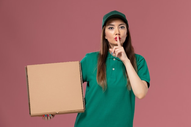 Corriere femminile di vista frontale in scatola di cibo della tenuta dell'uniforme verde sulla consegna uniforme di servizio di lavoro della parete rosa chiaro