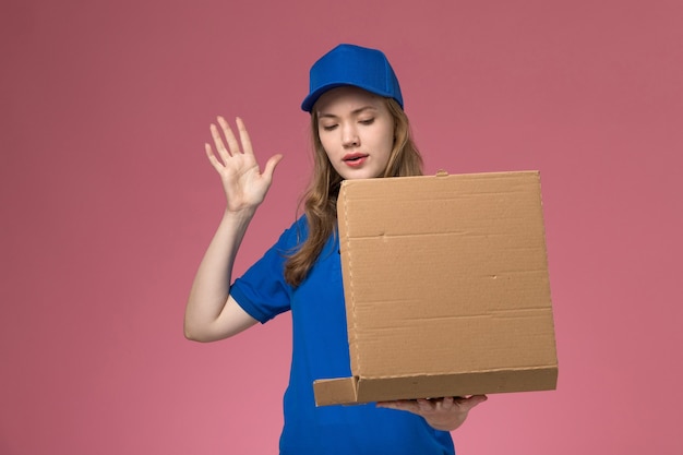 Corriere femminile di vista frontale in scatola di consegna del cibo della tenuta dell'uniforme blu che posa con esso sulla società dell'uniforme di servizio del lavoratore del fondo rosa