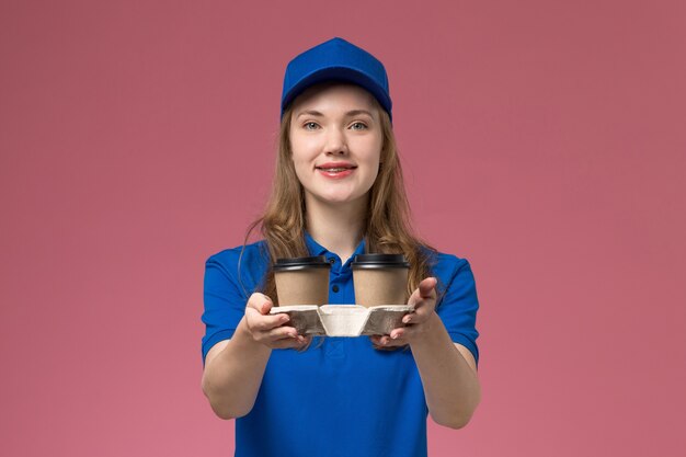 Corriere femminile di vista frontale in uniforme blu che consegna le tazze di caffè marroni sull'uniforme di servizio di scrivania rosa che consegna il lavoro dell'azienda