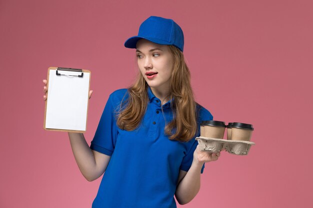 Corriere femminile di vista frontale in uniforme blu che tiene il blocchetto per appunti e le tazze di caffè di consegna sull'azienda dell'uniforme di servizio di scrittorio rosa chiaro