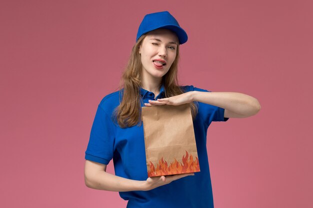 Corriere femminile di vista frontale in uniforme blu che tiene il pacchetto dell'alimento che mostra la sua linguetta sulla società dell'uniforme di servizio dell'operaio di lavoro dello scrittorio rosa