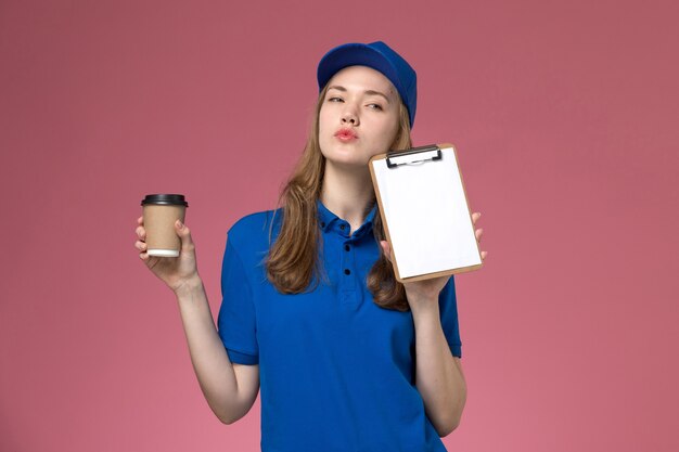 Corriere femminile di vista frontale in uniforme blu che tiene la tazza di caffè marrone con il blocchetto per appunti sull'uniforme di lavoro di servizio del fondo rosa chiaro che consegna azienda