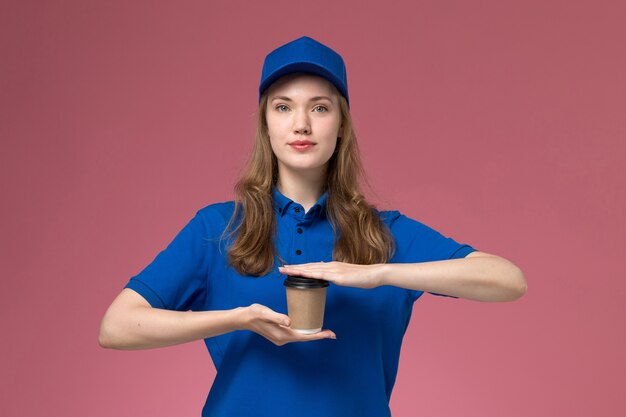 Corriere femminile di vista frontale in uniforme blu che tiene la tazza di caffè marrone sulla società di consegna del lavoro dell'uniforme di lavoro di servizio della scrivania rosa-chiaro