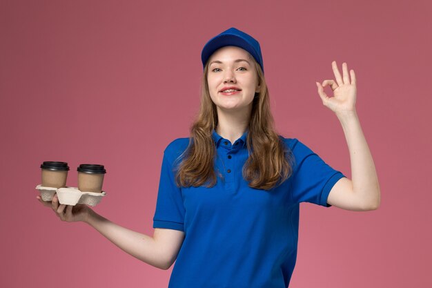 Corriere femminile di vista frontale in uniforme blu che tiene le tazze di caffè di consegna marroni che sorridono sull'uniforme di servizio del fondo rosa che consegna il lavoro dell'azienda