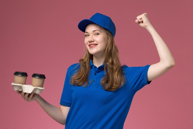 Corriere femminile di vista frontale in uniforme blu che tiene le tazze di caffè marroni che flettono con il sorriso sull'uniforme di servizio del fondo rosa che consegna il lavoro dell'azienda