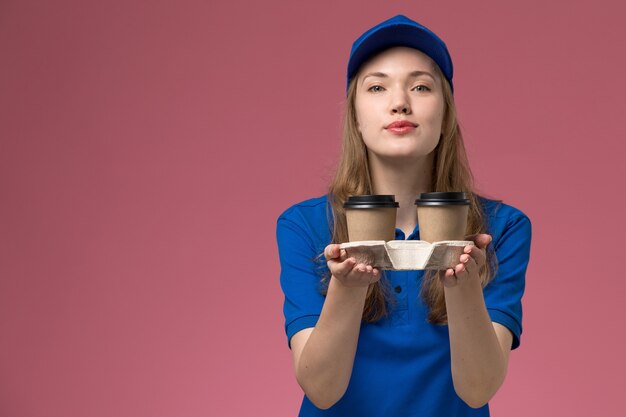 Corriere femminile di vista frontale in uniforme blu che tiene le tazze di caffè marroni che li consegna sull'uniforme di servizio del fondo rosa che consegna il lavoro dell'azienda