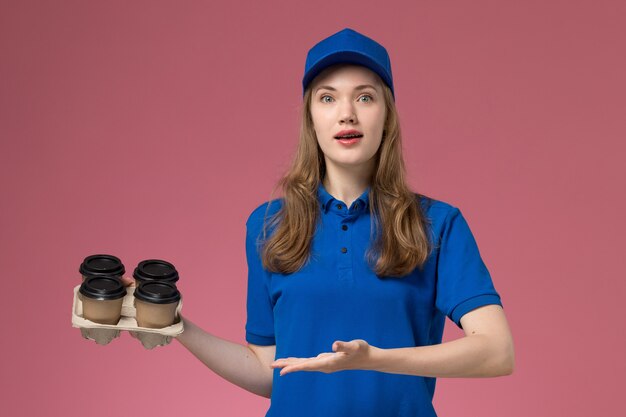 Corriere femminile di vista frontale in uniforme blu che tiene le tazze di caffè marroni di consegna sul lavoratore dell'azienda uniforme di servizio del fondo rosa