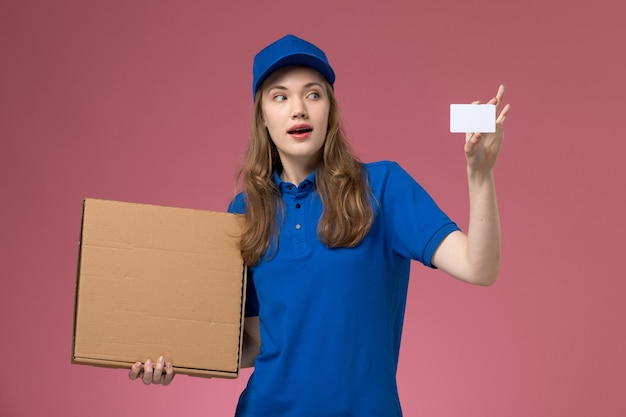 Corriere femminile di vista frontale in uniforme blu che tiene scatola di consegna del cibo e carta bianca sul lavoro aziendale uniforme di servizio scrivania rosa chiaro