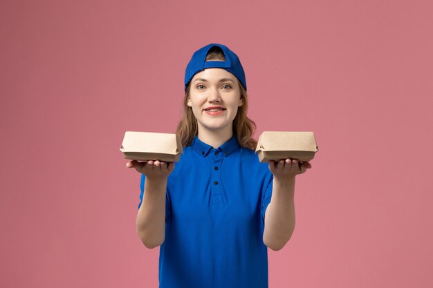 Corriere femminile di vista frontale in uniforme blu e mantello che tiene i piccoli pacchetti dell'alimento di consegna che sorridono sulla parete rosa, società di servizi dell'uniforme di lavoro di consegna