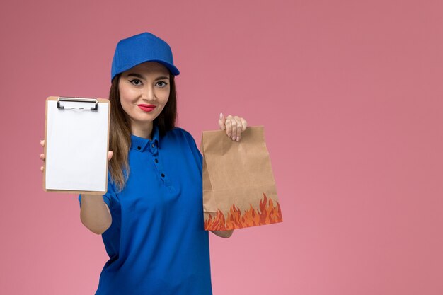 Corriere femminile di vista frontale in uniforme blu e mantello che tiene il blocco note con il pacchetto di cibo di carta sulla scrivania rosa