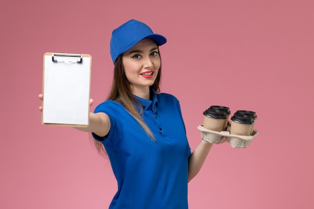 Corriere femminile di vista frontale in uniforme blu e mantello che tiene le tazze di caffè con il blocco note sulla donna rosa della parete