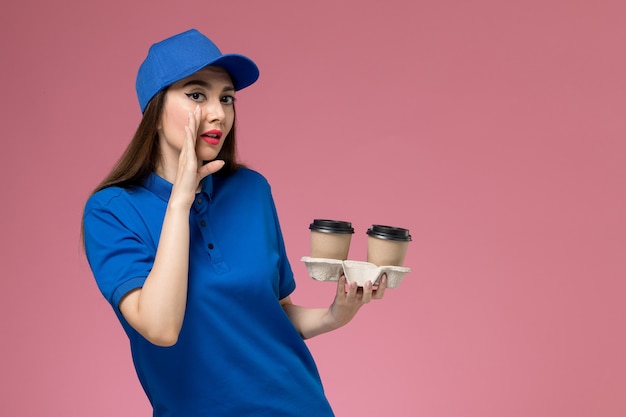 Corriere femminile di vista frontale in uniforme blu e mantello che tiene le tazze di caffè marroni di consegna che bisbigliano sulla parete rosa