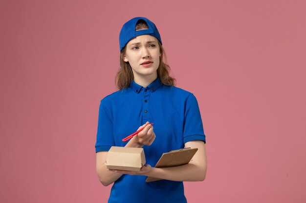 Corriere femminile di vista frontale in uniforme blu e mantello che tiene piccolo blocco note del pacchetto di cibo di consegna e scrittura sul muro rosa, lavoro dei dipendenti del servizio di consegna