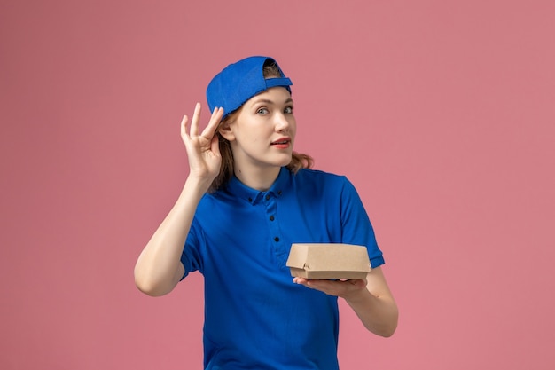 Corriere femminile di vista frontale in uniforme blu e mantello che tiene piccolo pacchetto di cibo di consegna cercando di sentire sul muro rosa, lavoro di società di servizi di consegna uniforme