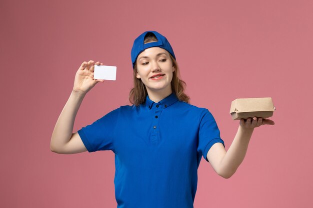 Corriere femminile di vista frontale in uniforme blu e mantello che tiene piccolo pacchetto di cibo di consegna con carta sulla parete rosa, impiegato di lavoro di servizio di consegna