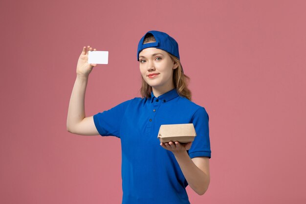 Corriere femminile di vista frontale in uniforme blu e mantello che tiene piccolo pacchetto di cibo di consegna con carta sulla parete rosa, impiegato di servizio di consegna