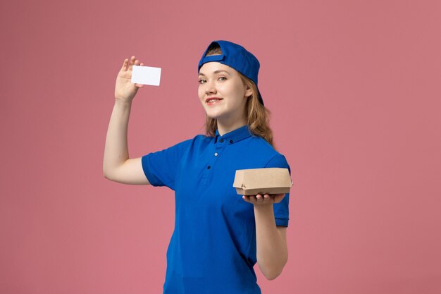 Corriere femminile di vista frontale in uniforme blu e mantello che tiene piccolo pacchetto di cibo di consegna con carta sulla parete rosa, impiegato di servizio di lavoro di consegna