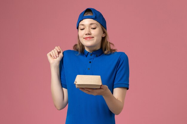 Corriere femminile di vista frontale in uniforme blu e mantello che tiene piccolo pacchetto di cibo di consegna sulla parete rosa chiaro, impiegato di servizio dell'operaio dell'uniforme di consegna